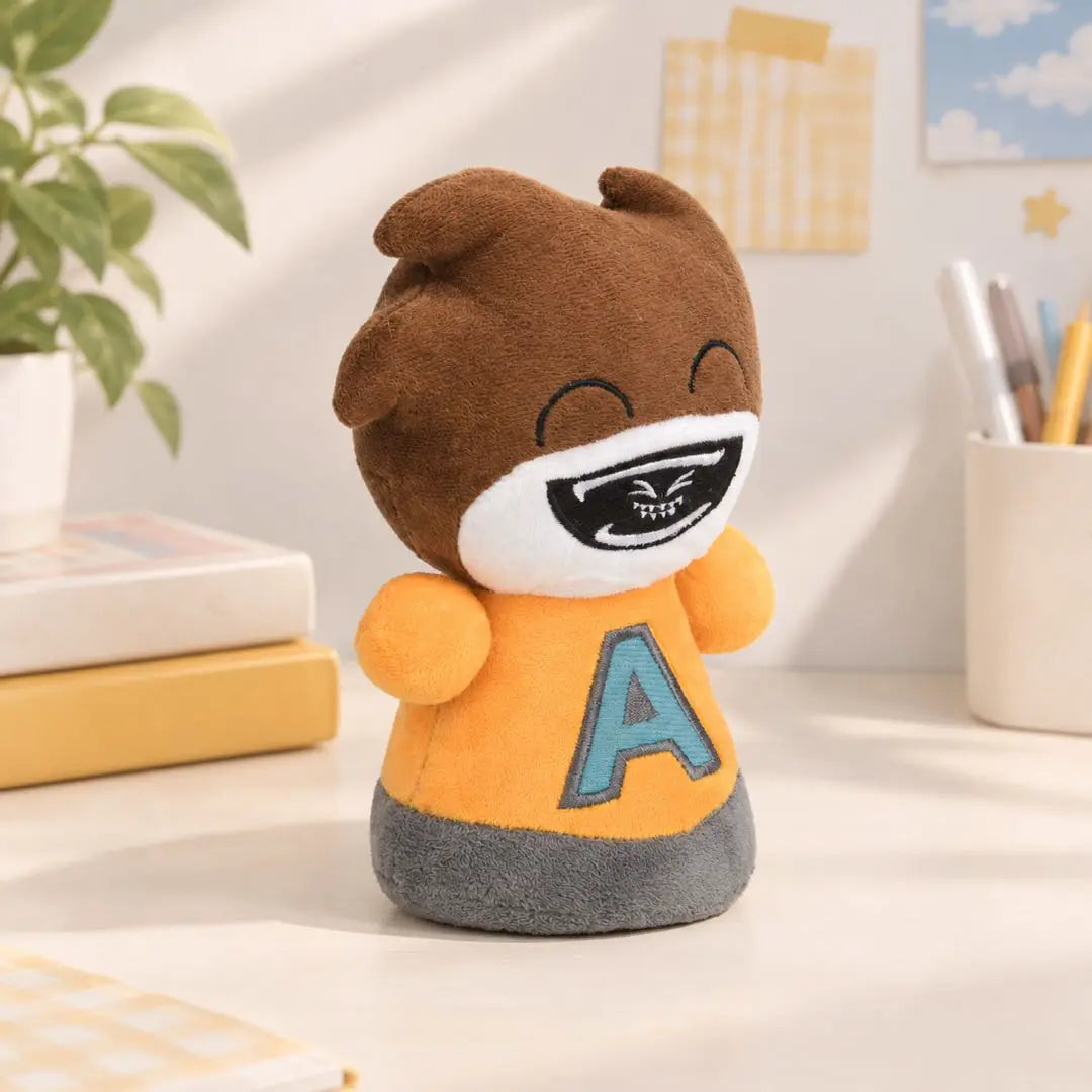 A plush toy character standing on a desk, featuring brown hair, a white face with a black mouth design, and an orange shirt with a blue letter 'A'. The background shows a softly lit room with a plant, books, and stationery.